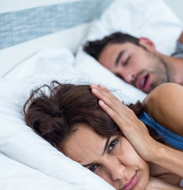 Irritated woman in bed covering her ears next to her snoring male partner 