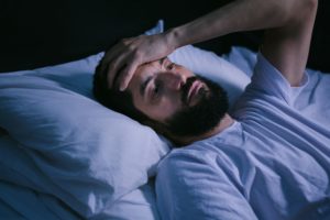 Man with black beard lying in bed with his hand to his forehead 