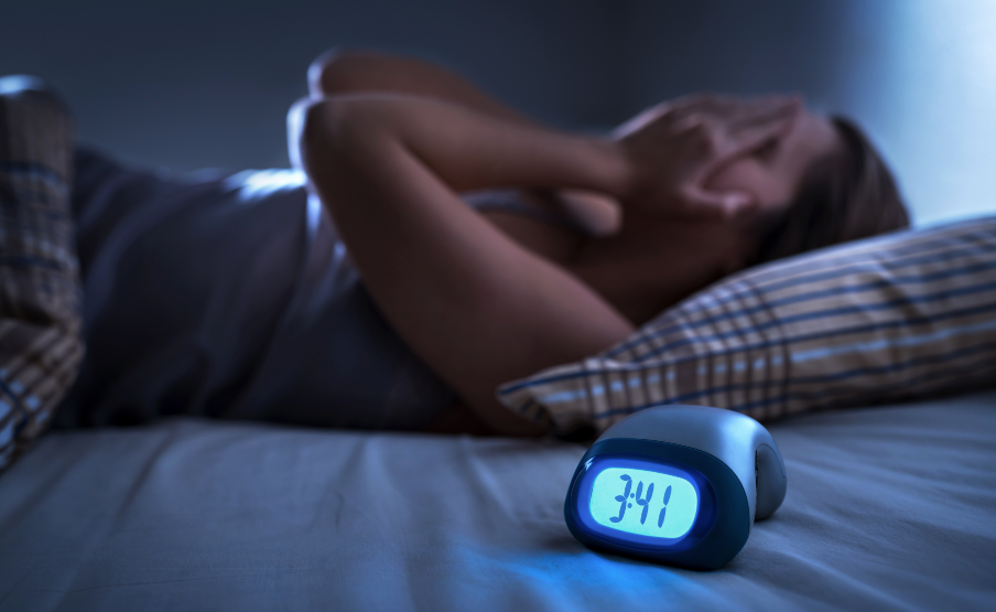 Woman with hands covering face, struggling to sleep, with alarm clock in the foreground
